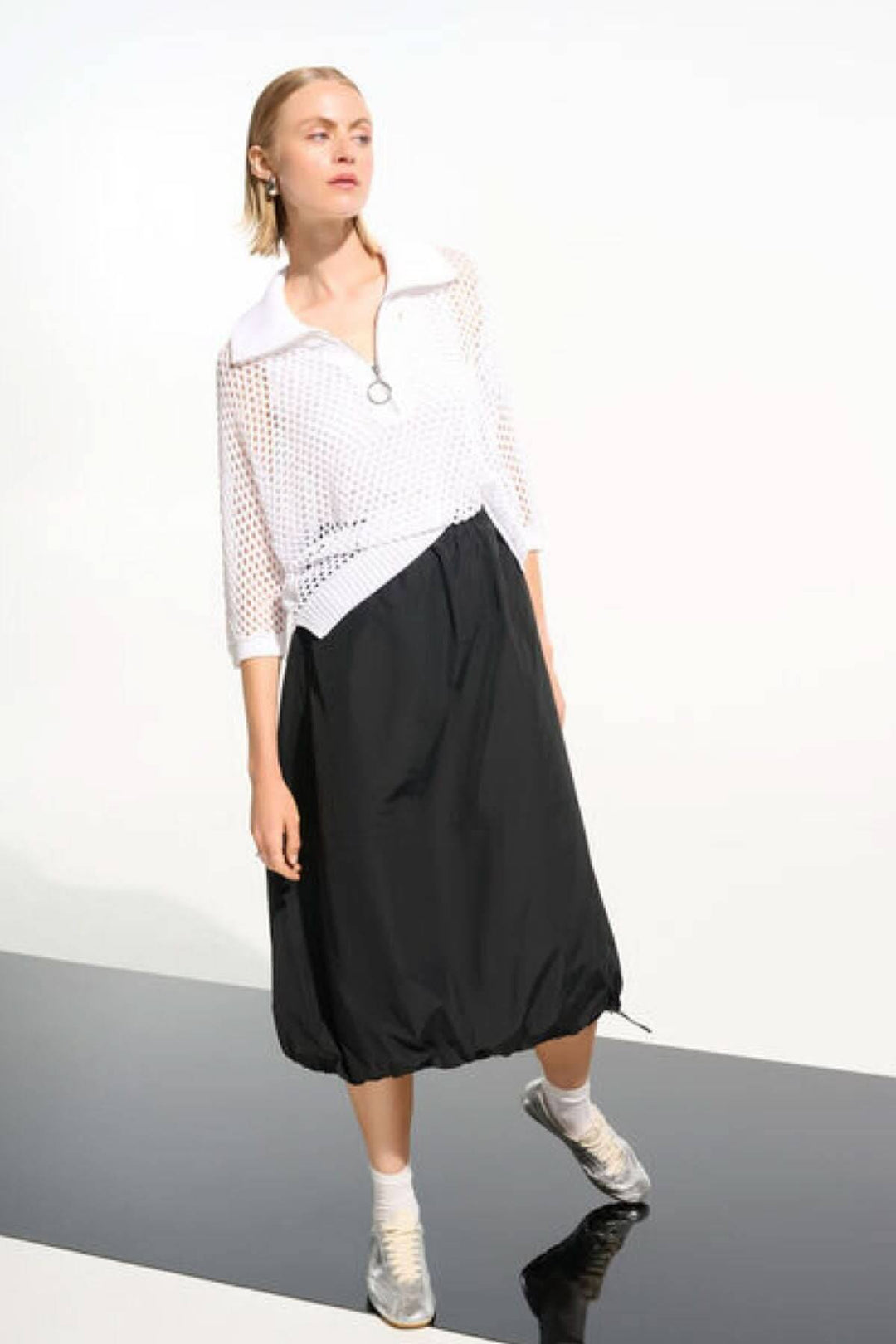 Modèle portant un chandail ajouré à col zippé avec manches trois-quarts et texture élégante, jupe noire ample, style moderne.
