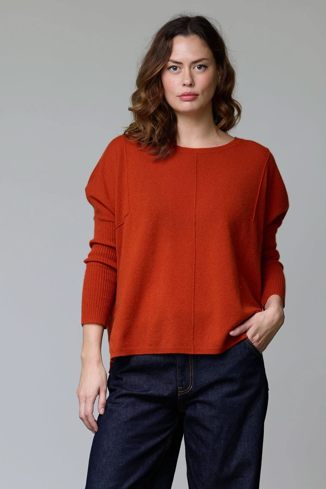 Femme portant un chandail ample à col rond, couleur orange, avec coutures apparentes et manches tombantes, style moderne et décontracté.