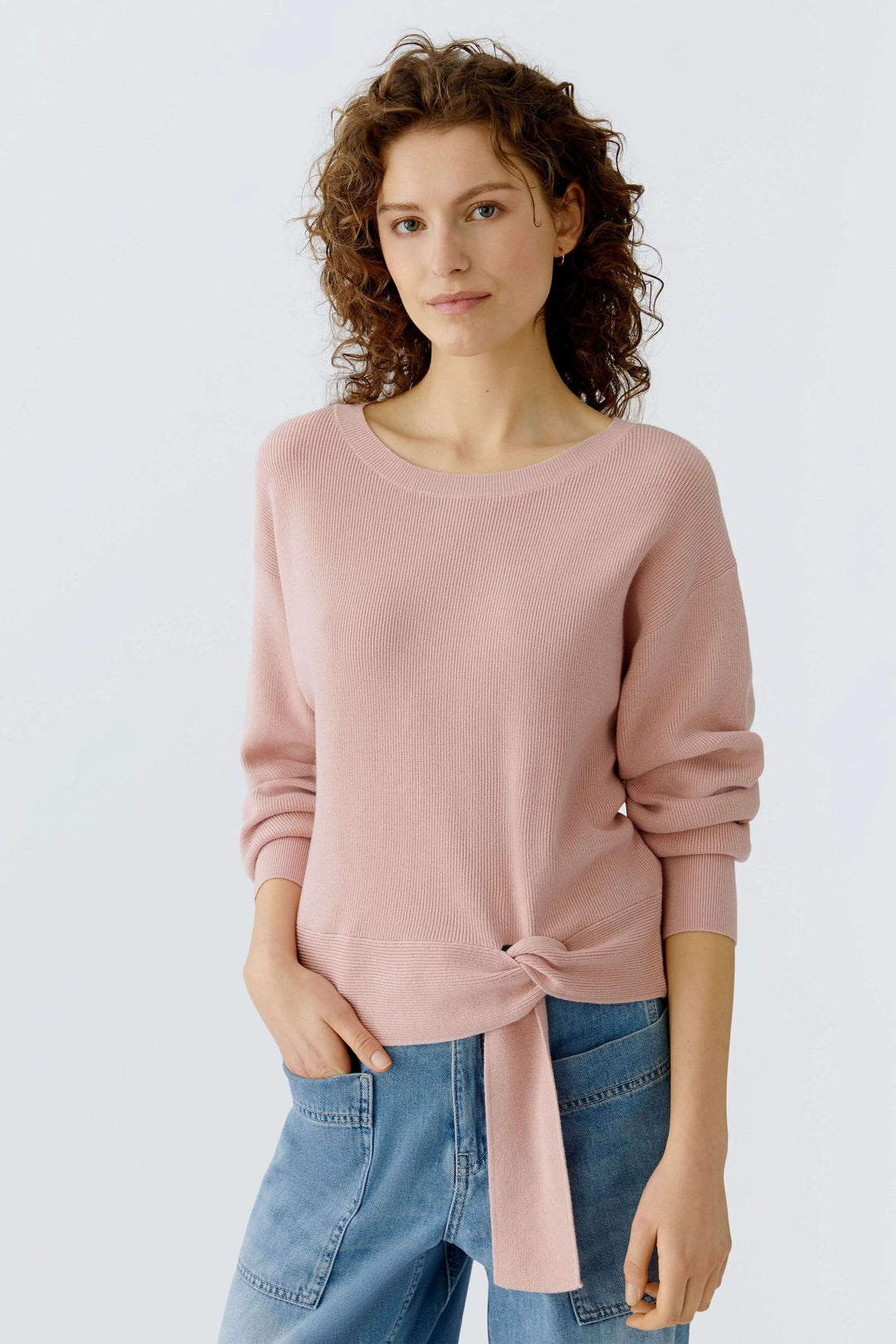 Femme portant un chandail rose avec un détail de nœud spécial à l'ourlet, style décontracté et moderne, idéal avec un jean.