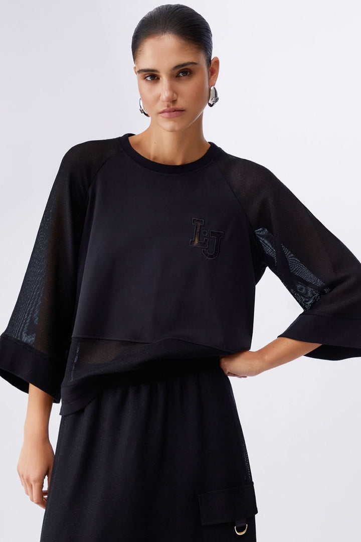 Femme portant un chandail court à manches larges, style moderne et décontracté, encolure ronde, couleur noire, avec écusson logo.