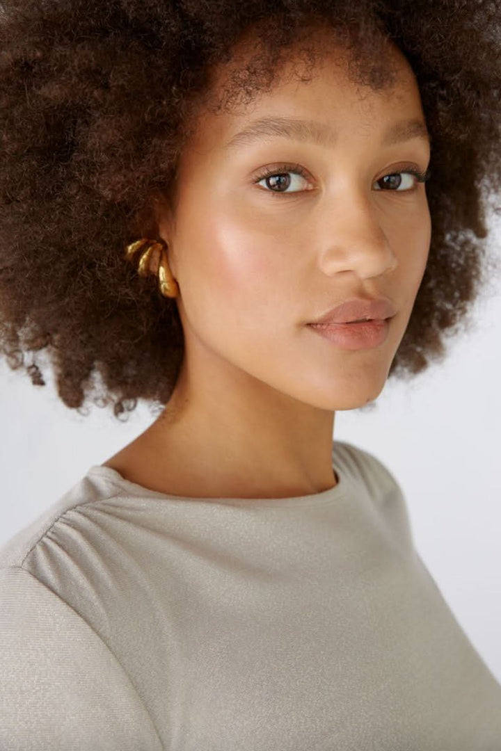 Femme avec cheveux crépus, portant un chandail brillant et des boucles d'oreilles dorées, regard confiant.
