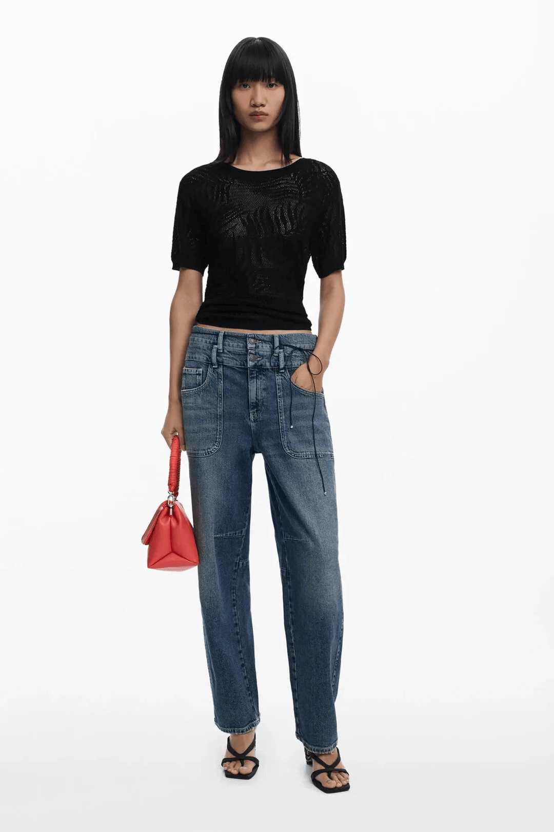 Femme portant un chandail en maille ajourée noire avec un pantalon denim, accessoire rouge dans la main.
