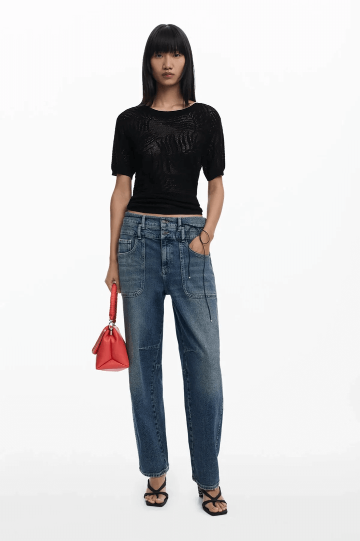 Femme portant un chandail en maille ajourée noire avec un pantalon denim, accessoire rouge dans la main.