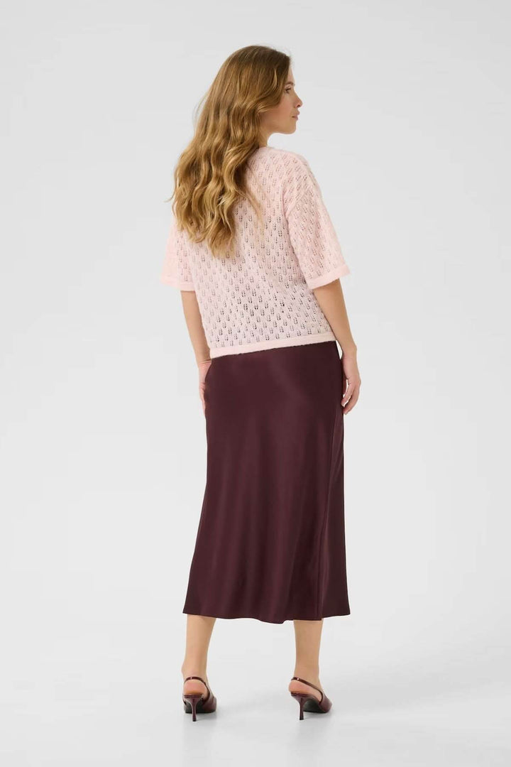 Femme vue de dos portant un chandail en tricot ajouré rose et une jupe longue bordeaux, sur un fond neutre.