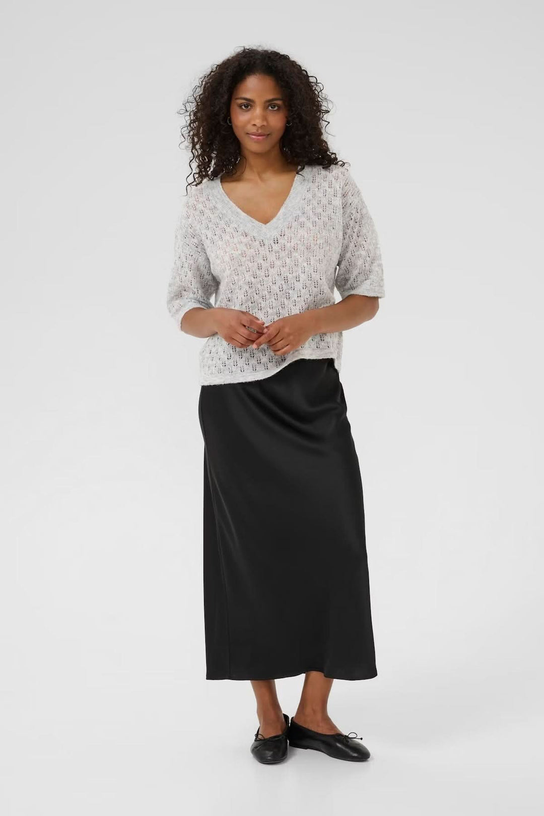Femme portant un chandail en tricot ajouré à col en V, jupe longue noire, look décontracté et féminin.