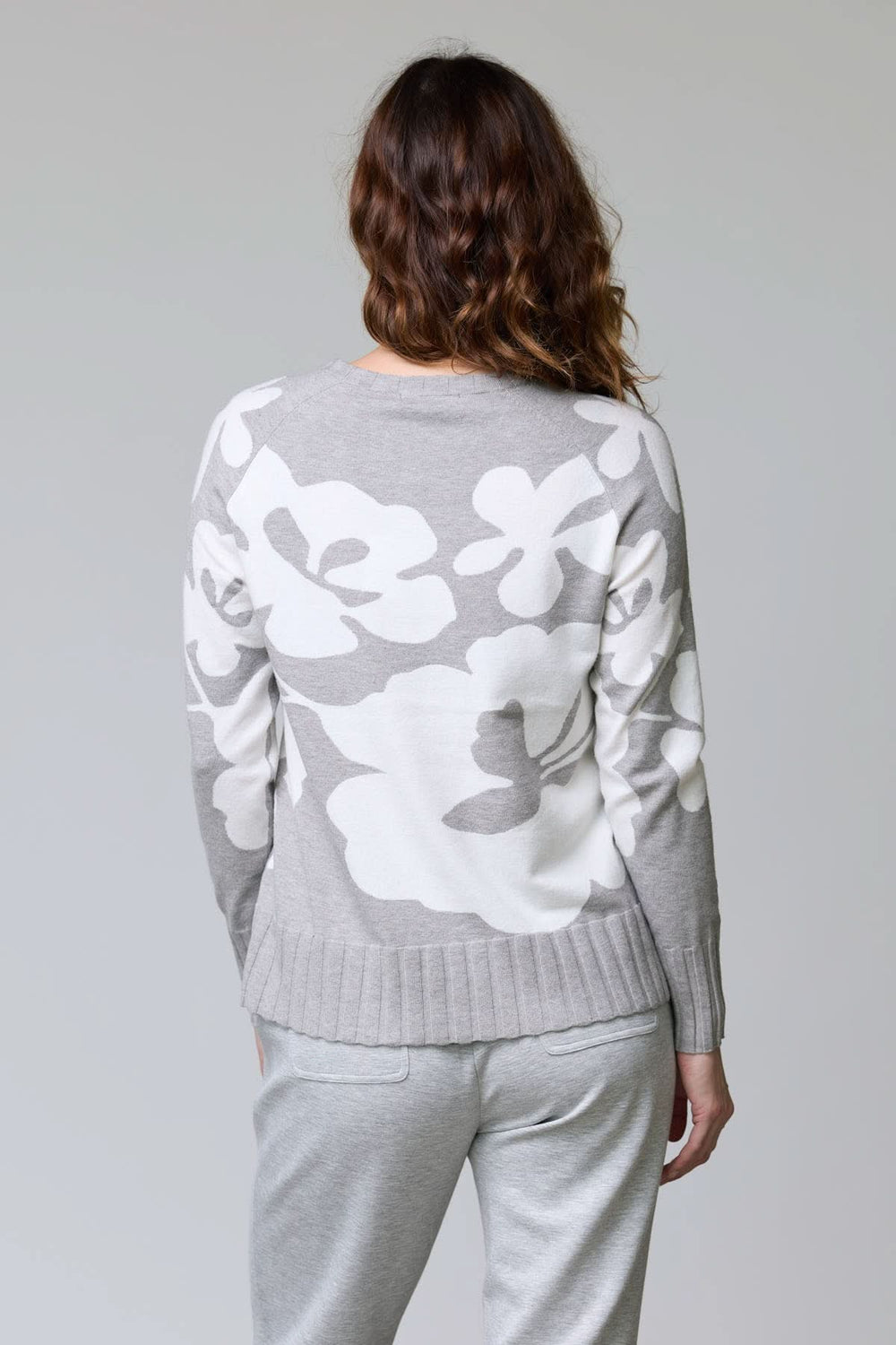 Femme portant un chandail en tricot à motif floral avec grandes fleurs blanches sur fond gris, vue de dos, coupe décontractée.