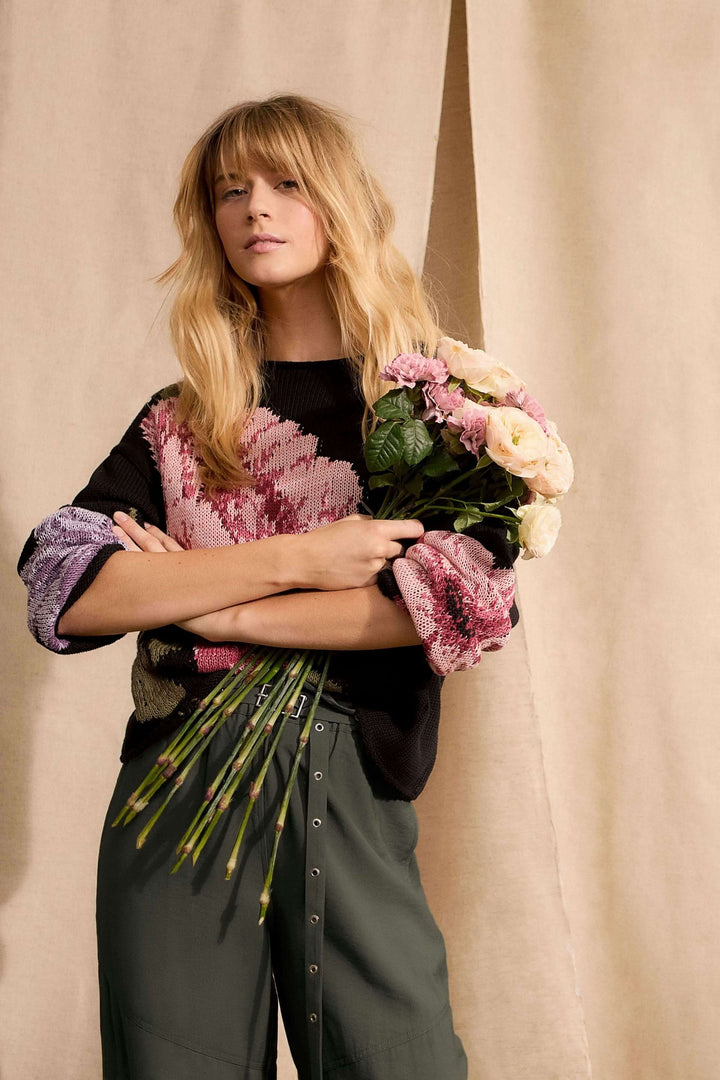 Femme portant un chandail imprimé floral avec un grand motif artistique, coupe décontractée et détails ajourés, tenant des fleurs.