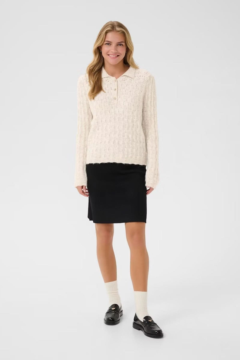 Femme souriante portant un chandail à maille torsadée crème avec col polo et jupe noire élégante.