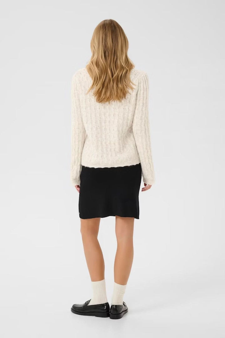 Femme vue de dos portant un chandail à maille torsadée beige avec une jupe noire et des chaussettes blanches.