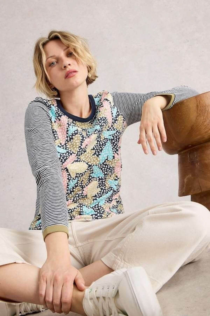 Femme portant un chandail à motifs graphiques avec imprimé floral et manches longues rayées, assise de manière décontractée.
