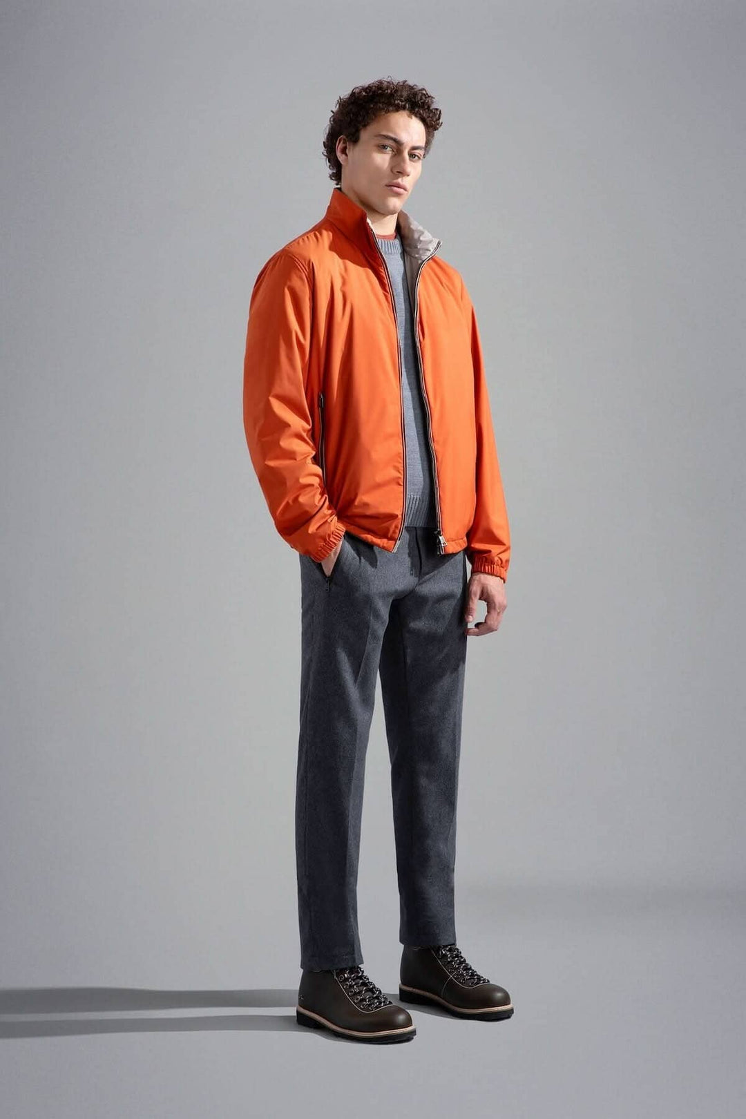 Homme en veste orange, pantalon gris et chaussures noires, posant dans un studio minimaliste.