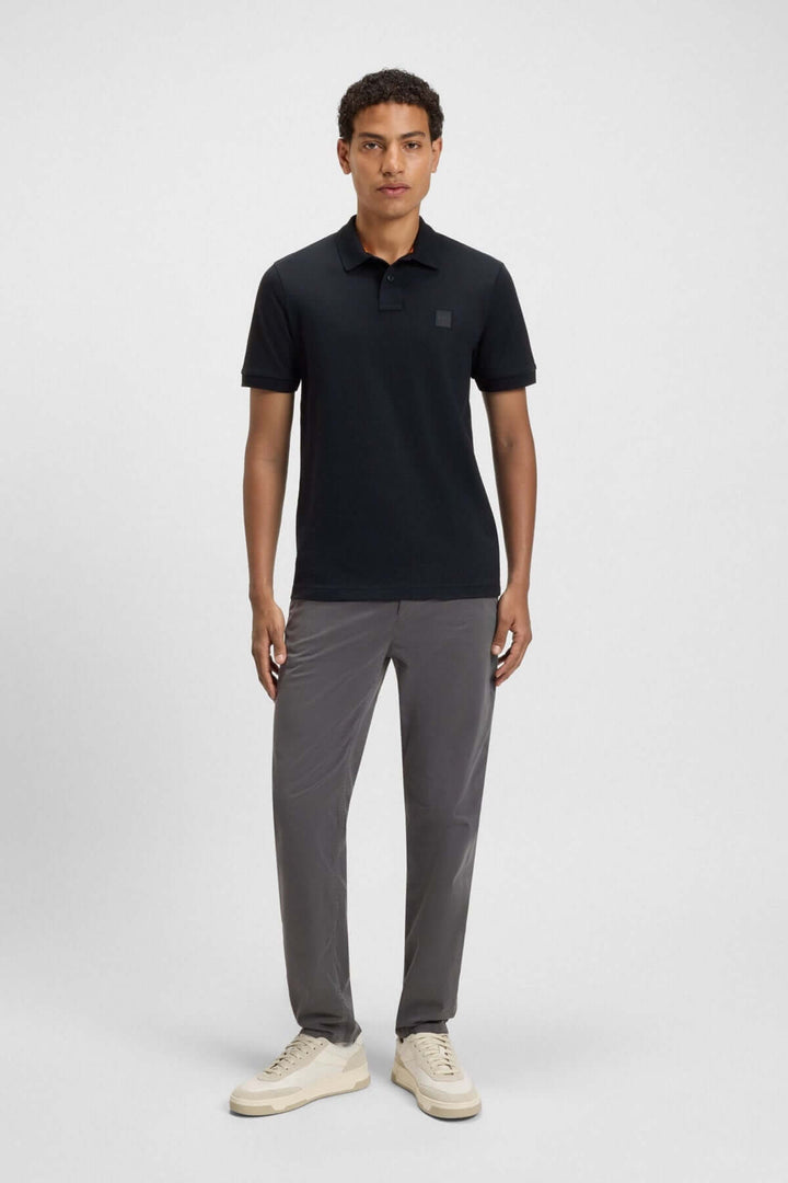 Homme en polo noir à logo Boss, pantalon gris, style décontracté et chic.