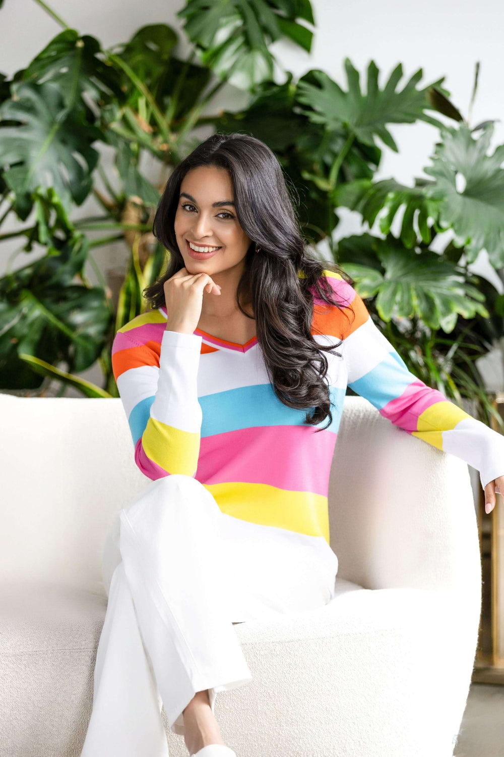 Femme portant un chandail à rayures multicolores avec encolure en V, assise sur un canapé beige, devant des plantes vertes.