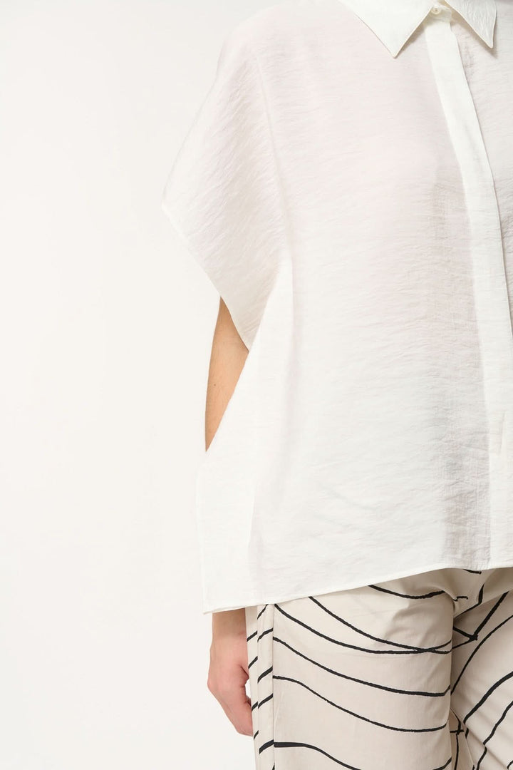 Légère chemise blanche à coupe ample, col classique et manches courtes, portée avec un pantalon à rayures.