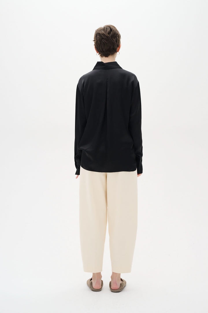 Chemise Pauline satinée noire vue de dos, portée avec un pantalon ample beige, offrant un style chic décontracté.