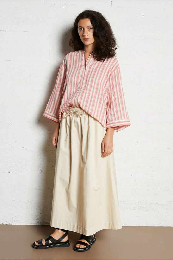 Femme portant une chemise design épuré rose à rayures avec une jupe beige, tenue moderne et élégante.