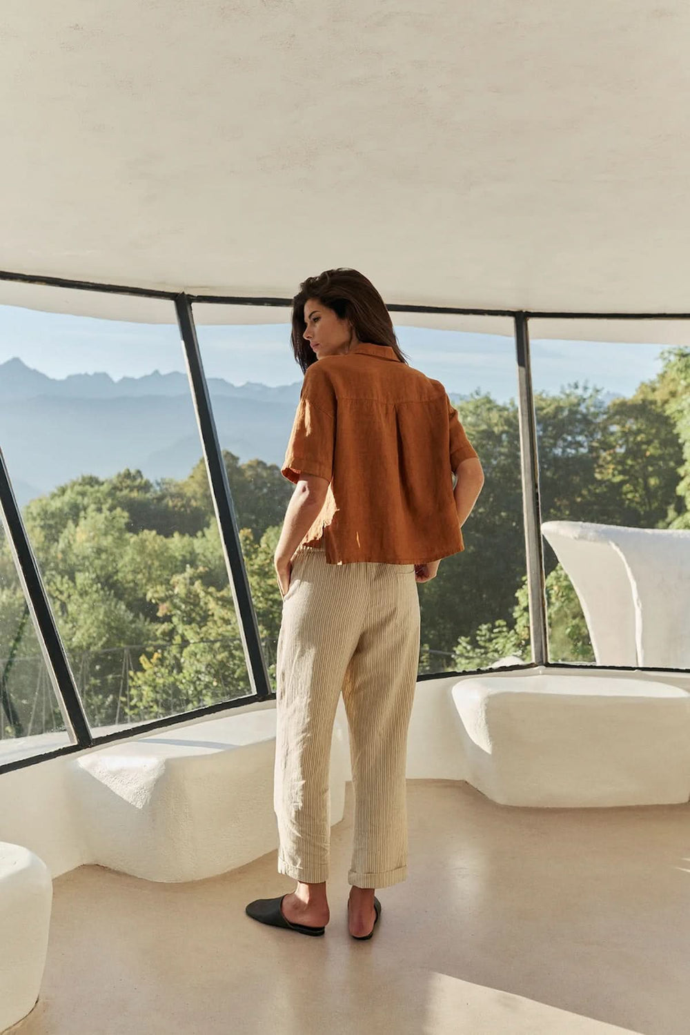Femme portant une chemise en lin à coupe décontractée dans un intérieur lumineux avec vue sur les montagnes et la forêt.