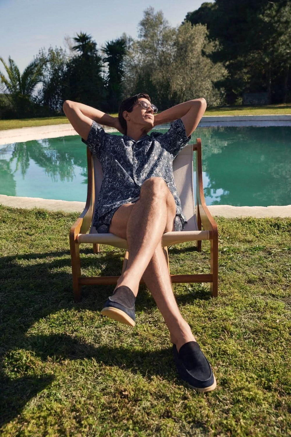 Homme détendu en chemise à imprimé tropical, assis sur une chaise près d'une piscine en plein air.