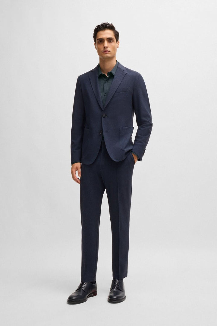 Homme en costume moderne bleu marine avec chemise verte, chaussures en cuir, pose élégante sur fond neutre.