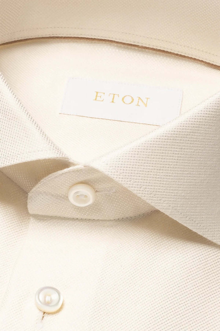 Détail d'une chemise Eton, en coton, avec col classique et boutons blancs sur fond beige.