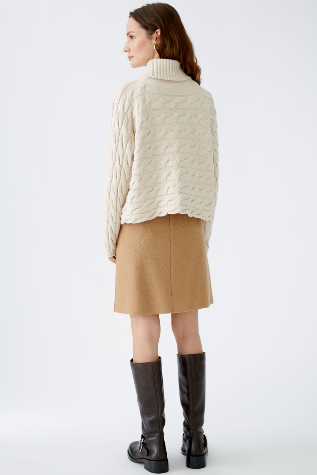 Femme vue de dos portant un col roulé en tricot torsadé avec jupe beige et bottes noires.
