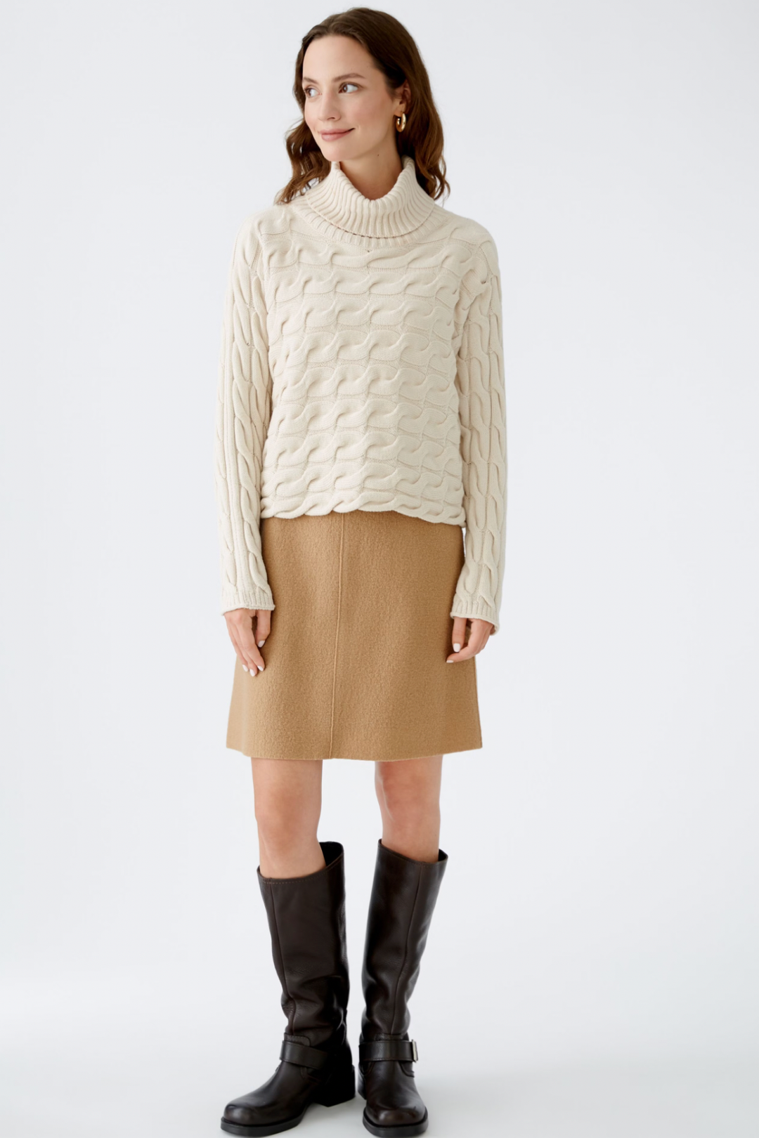 Femme en col roulé en tricot torsadé crémeux avec jupe beige et bottes noires, look élégant pour l'hiver.