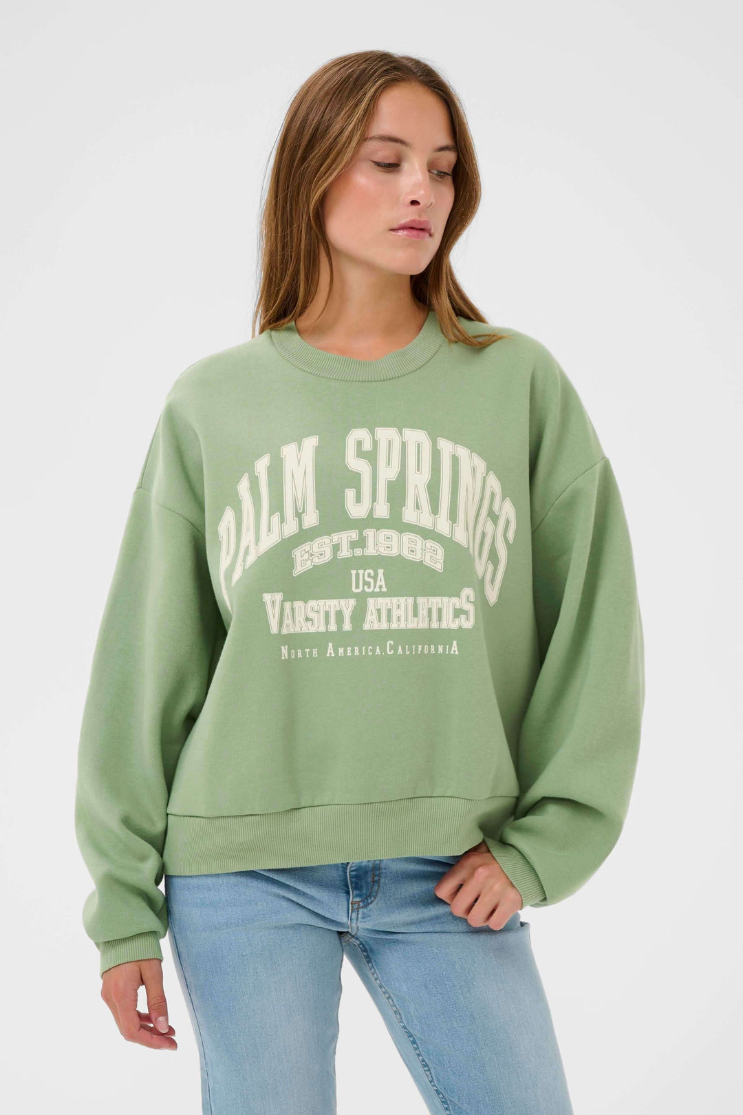 Femme portant un coton ouaté vert "Coton ouaté Sussi" au style universitaire avec motif imprimé "Palm Springs" et jean décontracté.