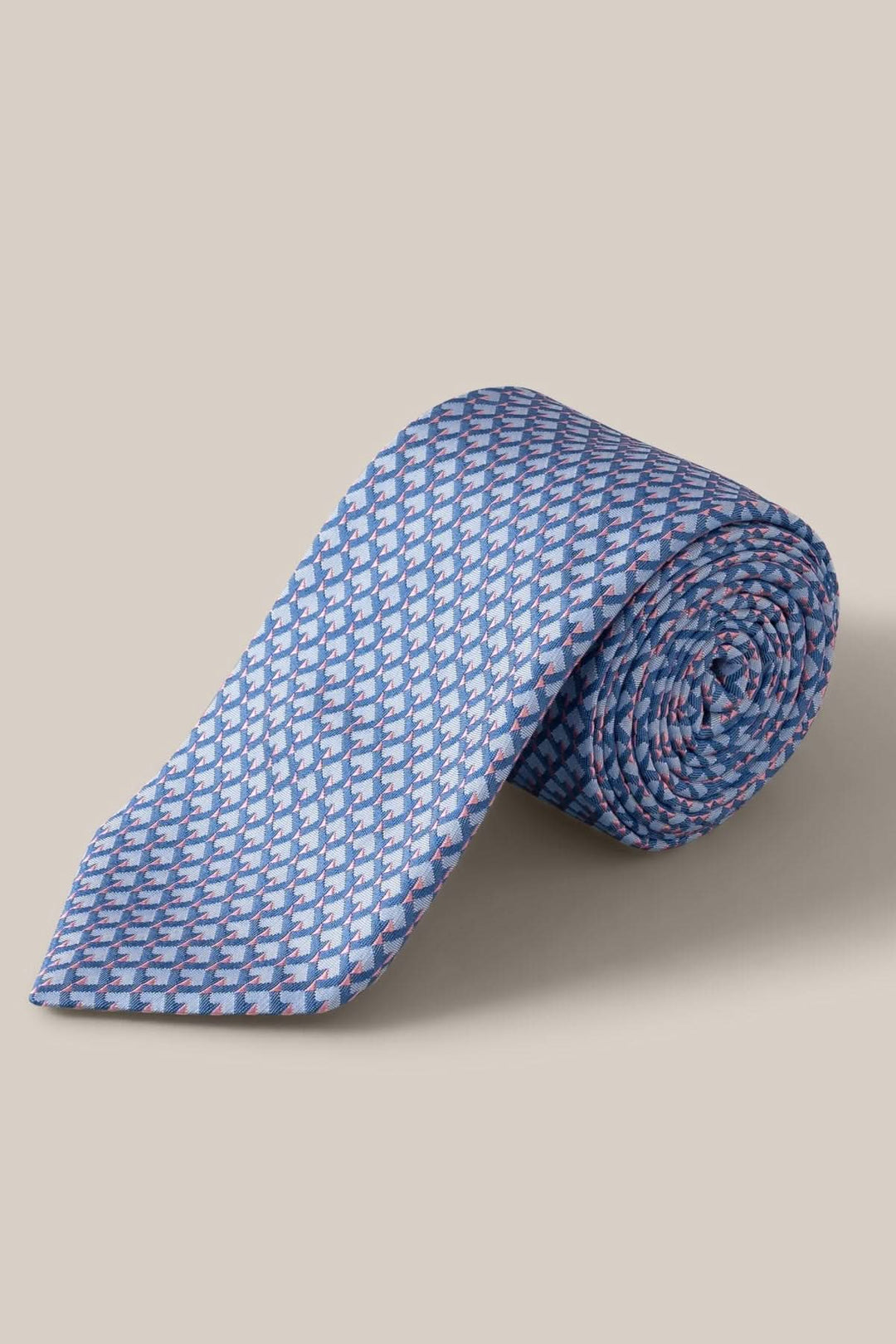 Cravate en soie à motif géométrique en nuances de bleu, gris et rose pâle, roulée sur une surface beige.