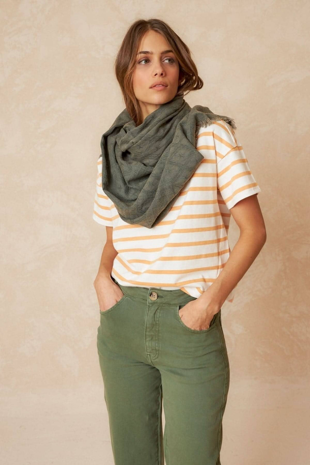 Foulard à motifs porté autour du cou par une femme en t-shirt rayé et pantalon vert, avec un fond neutre.