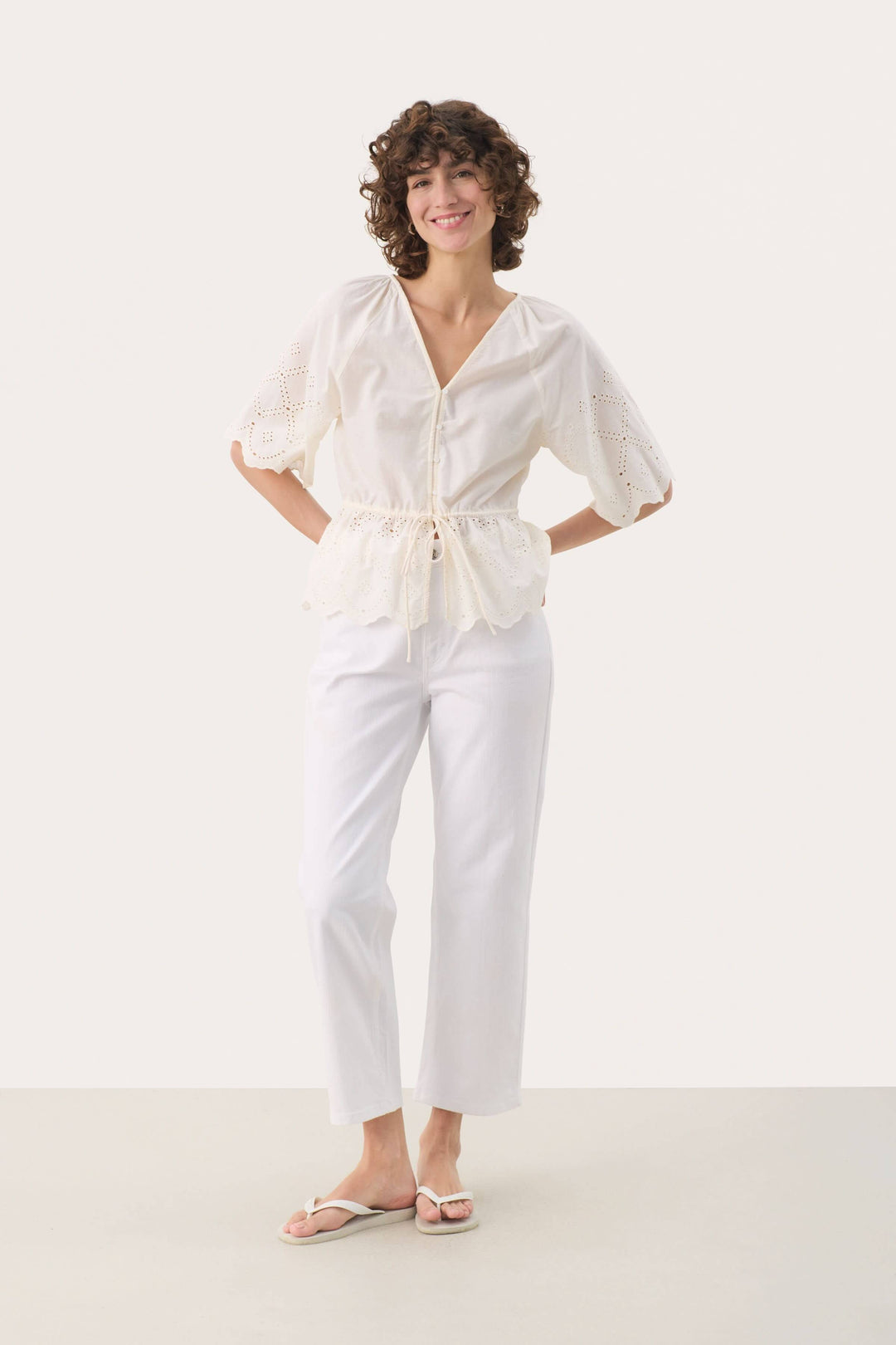 Femme souriante en blouse en coton blanc avec encolure en V et manches amples, portant un pantalon blanc.