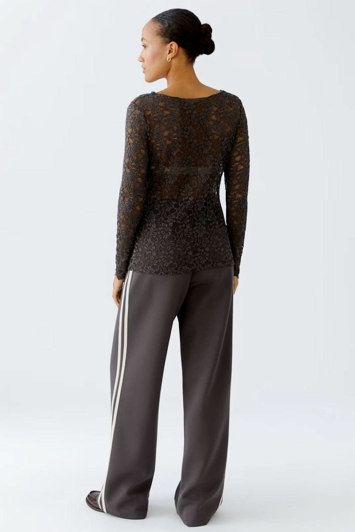 Femme portant un haut en dentelle florale noir opaque et élégant, manches longues, avec un col bateau, associé à un pantalon chic.