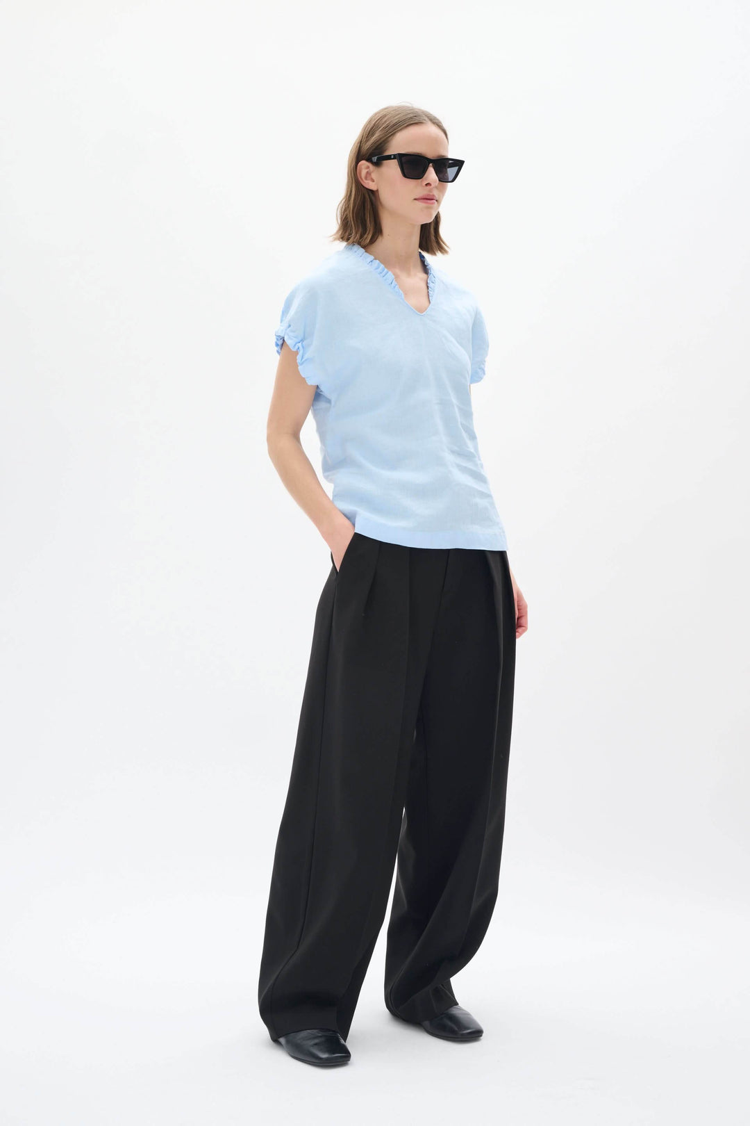 Femme portant un haut en lin bleu clair avec un col en V, lunettes de soleil et pantalon noir ample.