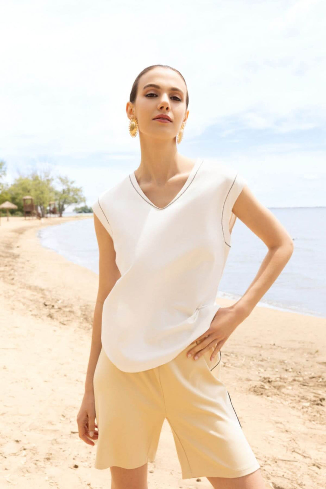 Femme portant un haut fluide col en V blanc sur une plage ensoleillée, style moderne et élégant.