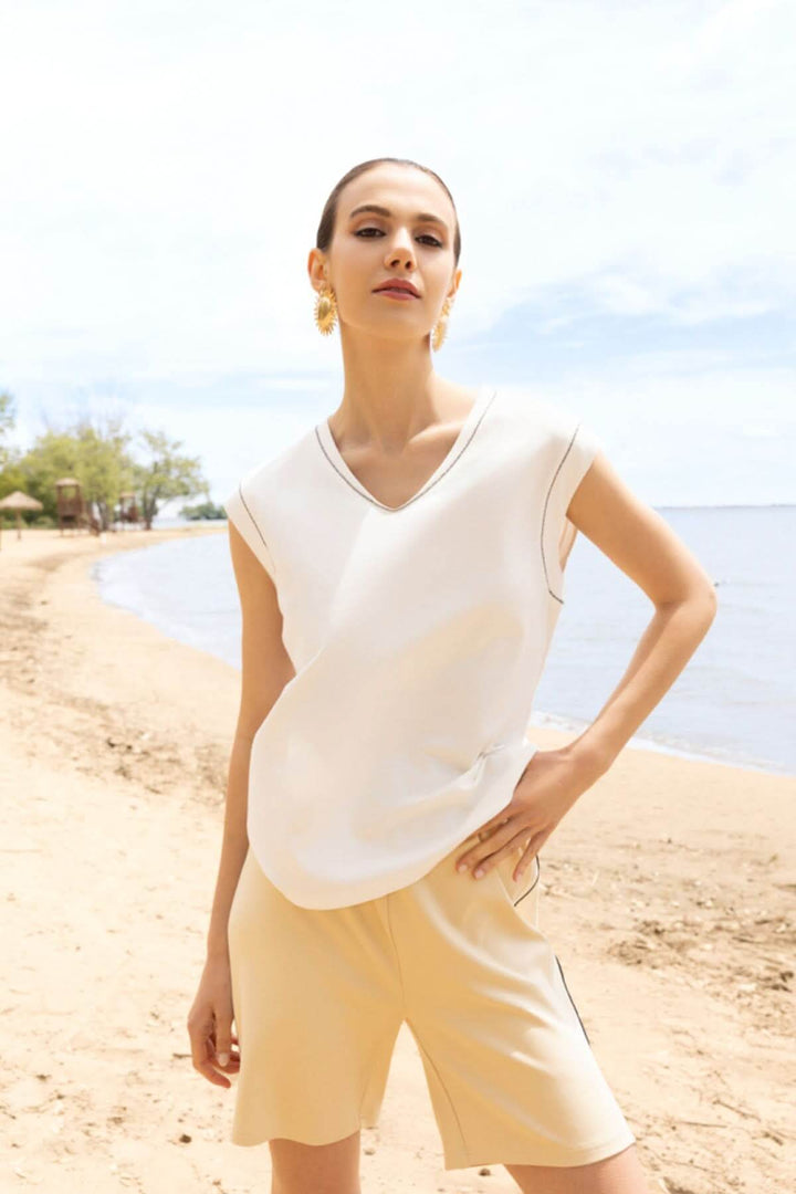 Femme portant un haut fluide col en V blanc sur une plage ensoleillée, style moderne et élégant.