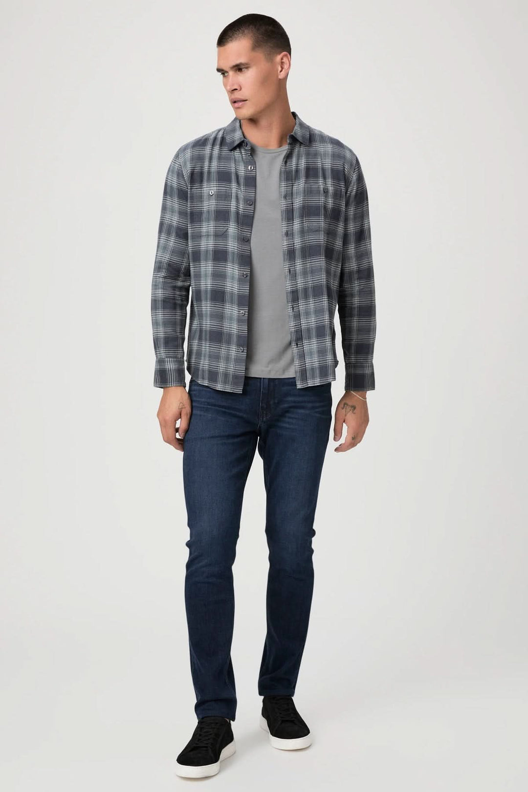Homme portant une chemise à carreaux grise et un t-shirt, avec un jean ajusté bleu foncé et des baskets noires.