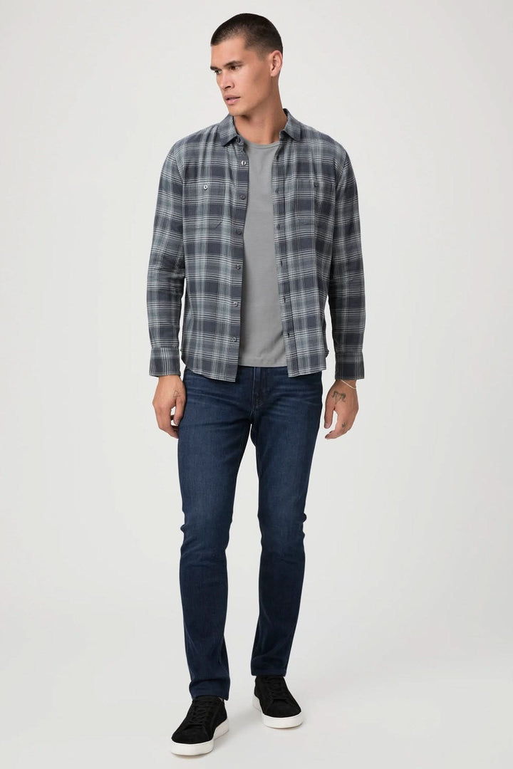 Homme portant une chemise à carreaux grise et un t-shirt, avec un jean ajusté bleu foncé et des baskets noires.