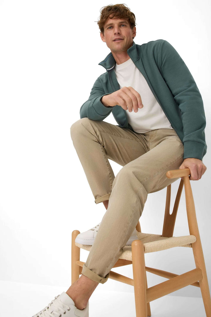 Homme assis sur une chaise, portant un gilet bleu moderne et un T-shirt blanc, jeans clairs, style décontracté.