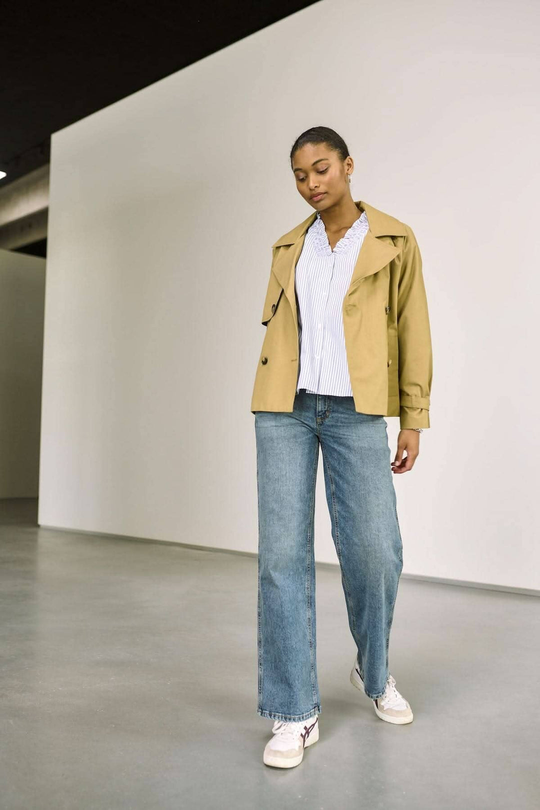 Femme portant un trench-beige avec un jean droit et des baskets blanches, look décontracté moderne.