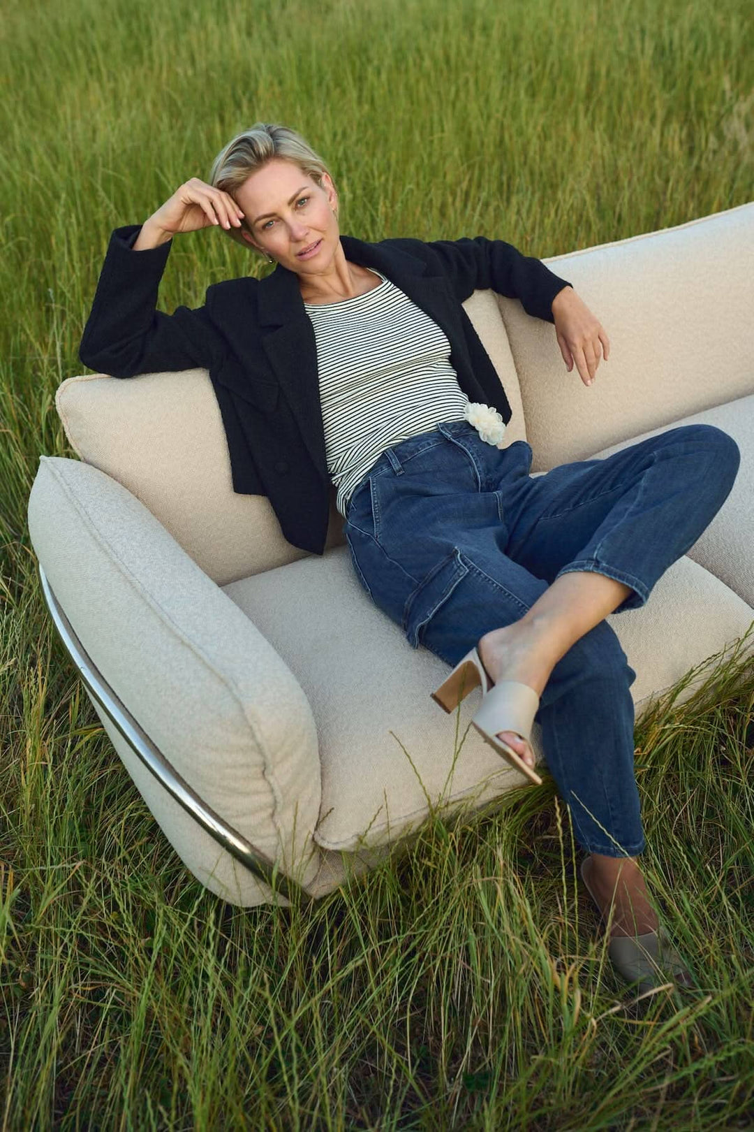 Femme élégante posant sur un canapé moderne dans un champ verdoyant, portant un haut rayé et un blazer noir.