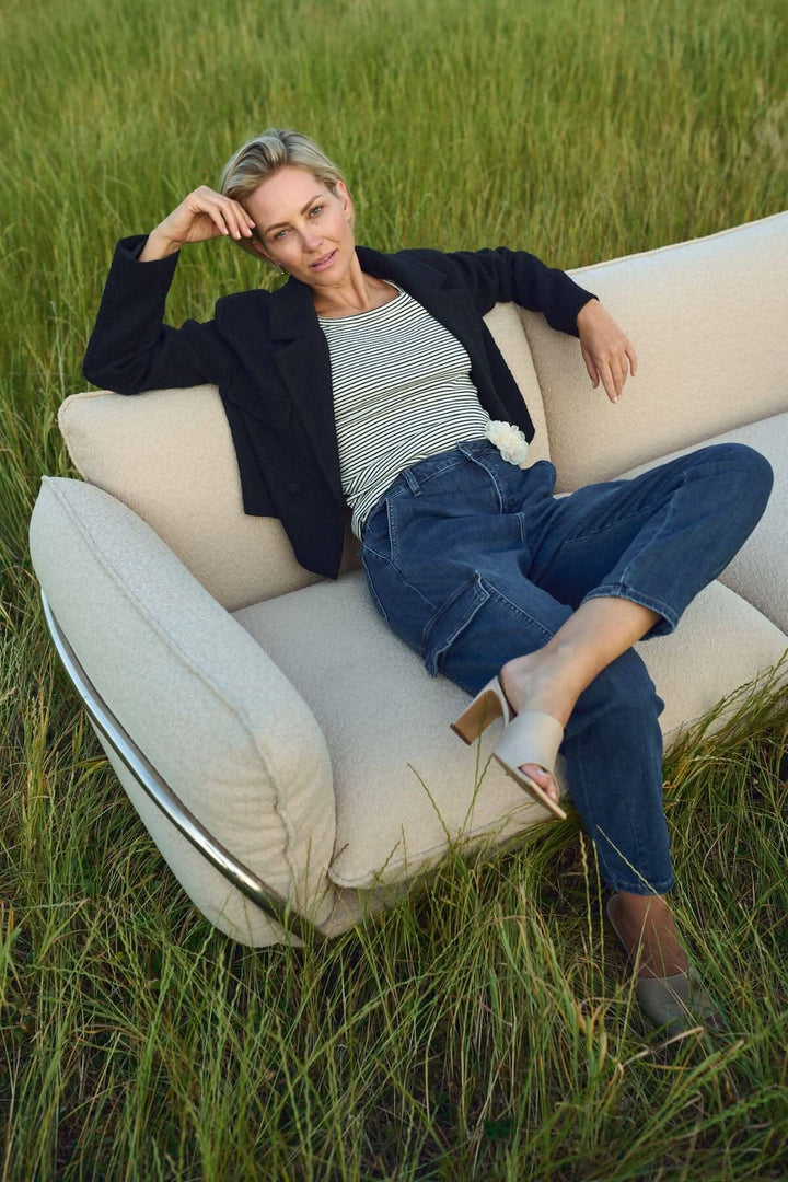 Femme élégante posant sur un canapé moderne dans un champ verdoyant, portant un haut rayé et un blazer noir.