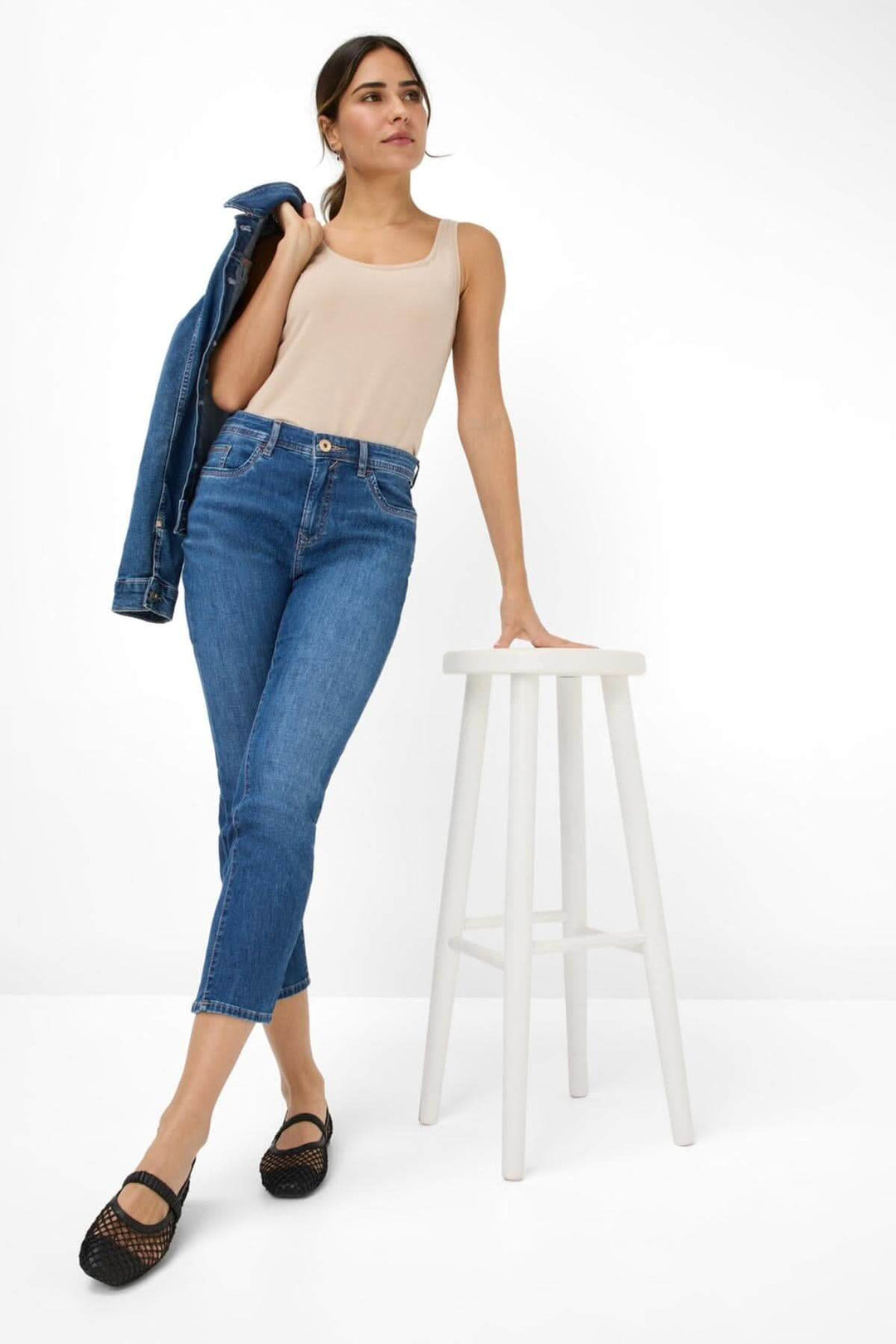 Femme portant le jean MARY S 6/8, combiné avec un débardeur beige et une veste, posant sur une tabouret blanc.
