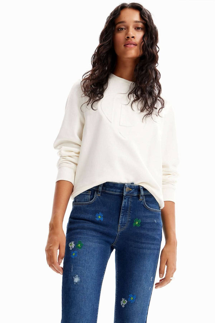 Femme portant un sweat-shirt blanc et un jean skinny bleu avec broderies florales et déchirures.