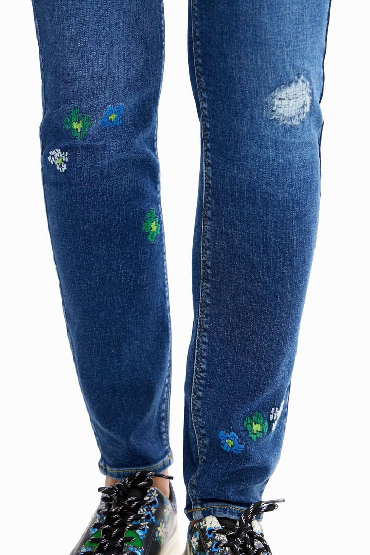 Jean skinny brodé avec déchirures et fleurs colorées, effet push-up, sur fond bleu.
