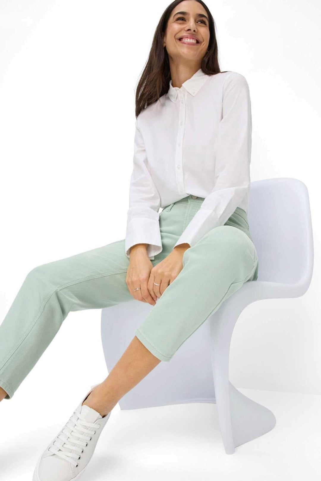 Femme souriante portant une chemise blanche et un pantalon vert, assise sur une chaise moderne.