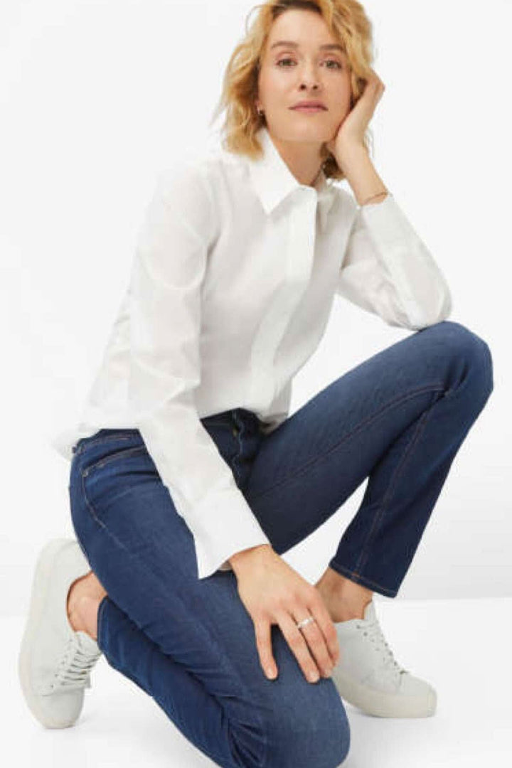 Femme en chemise blanche et jeans skinny, posant à genoux avec des baskets blanches, style moderne.