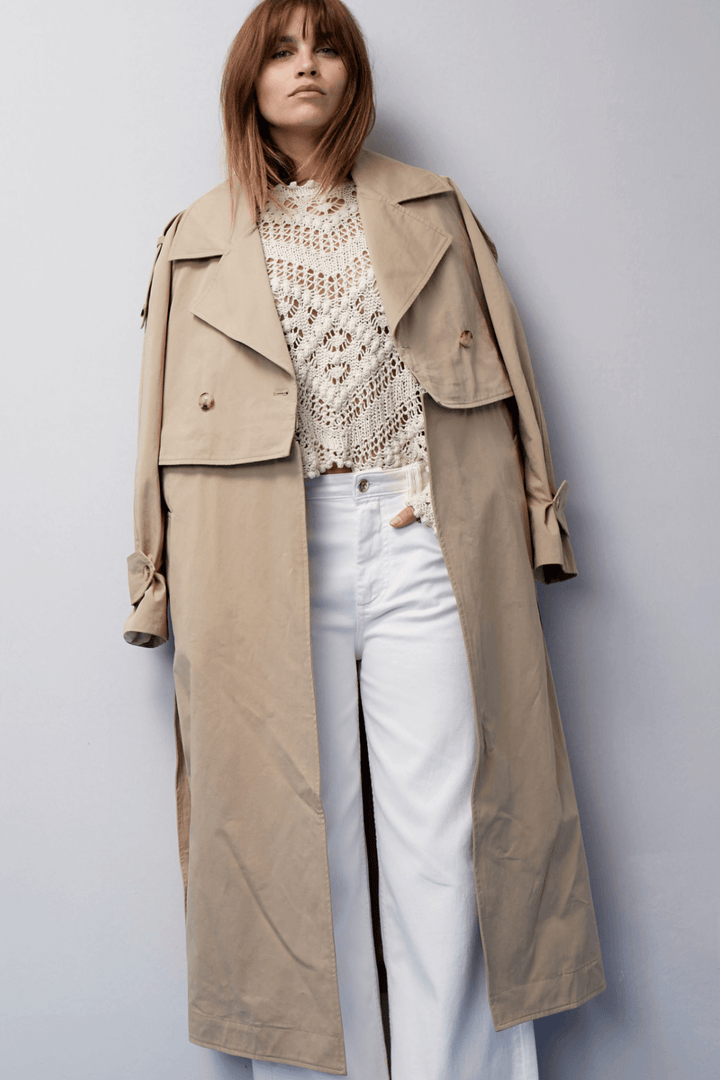 Femme portant un trench beige sur un haut en crochet, assorti à des jeans blancs modernes.