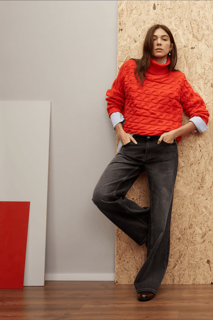Femme dans un pull rouge avec jeans évasés, posant contre un mur en bois, style décontracté et élégant.