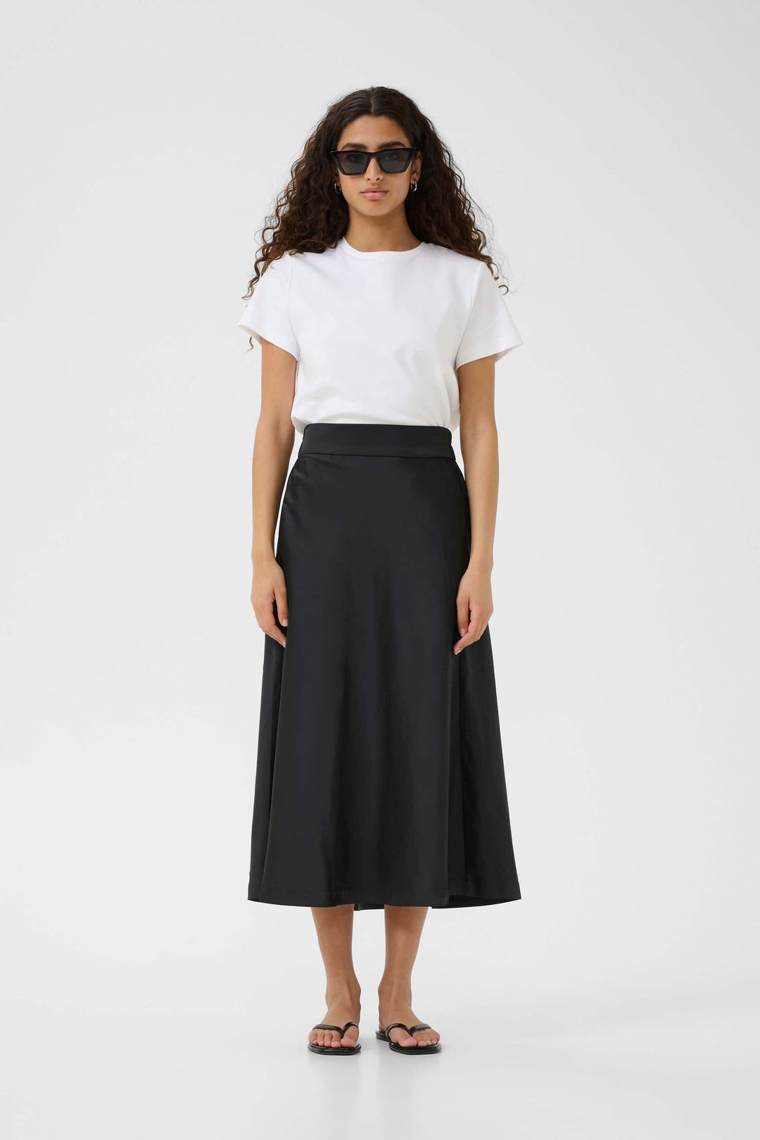 Femme portant une jupe midi noire fluide avec un t-shirt blanc et des sandales, look minimaliste élégant.