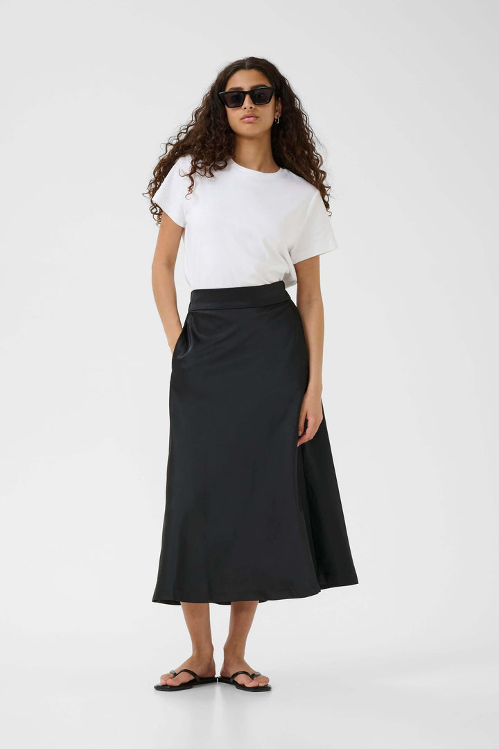 Femme en jupe midi fluide noire avec t-shirt blanc, style minimaliste et élégant, porte des lunettes de soleil.