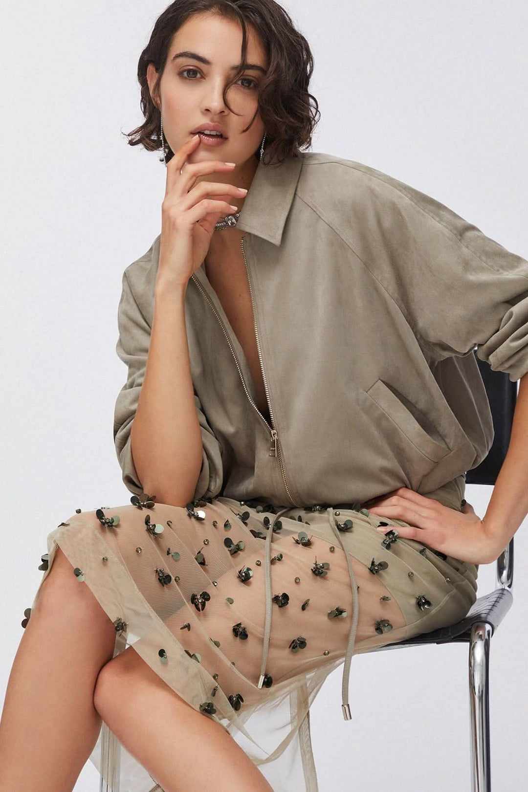 Jupe en maille transparente ornée avec détails appliqués sur fond clair, portée avec une veste taupe, créant une allure moderne.